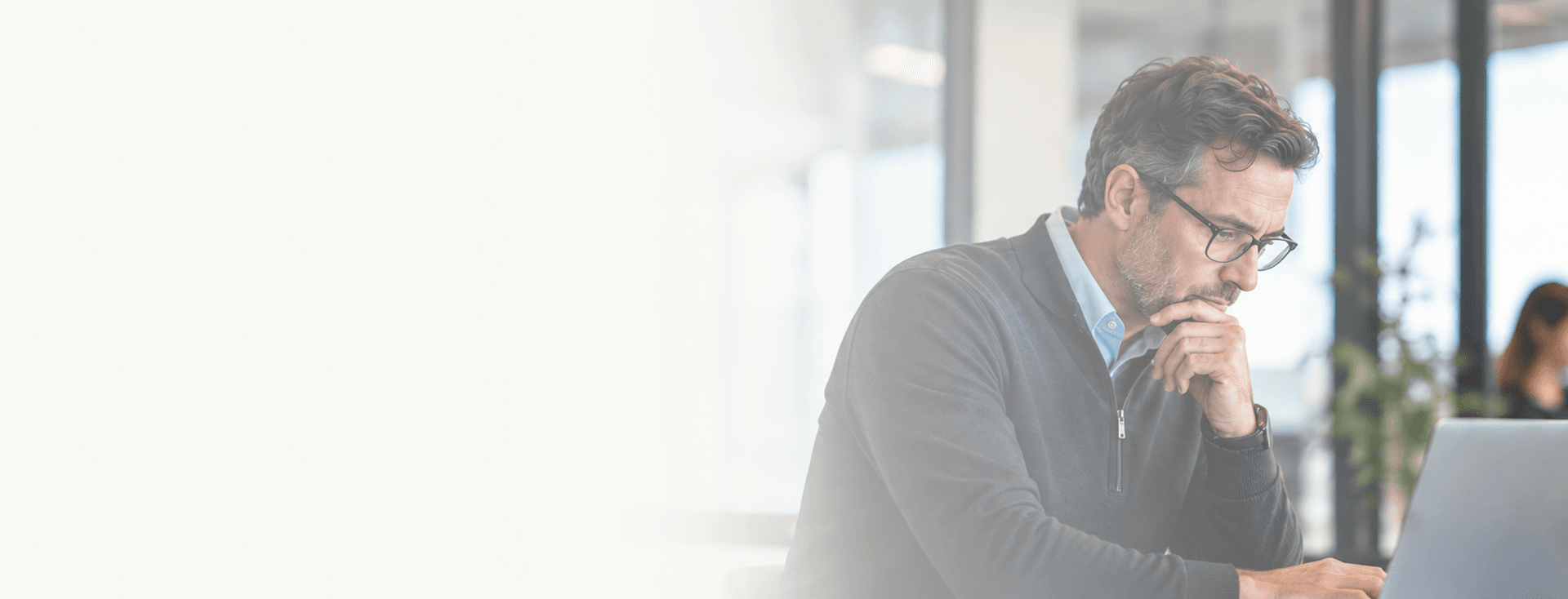 Man in glasses resting his chin on his hand while reading a laptop in a bright open-plan office