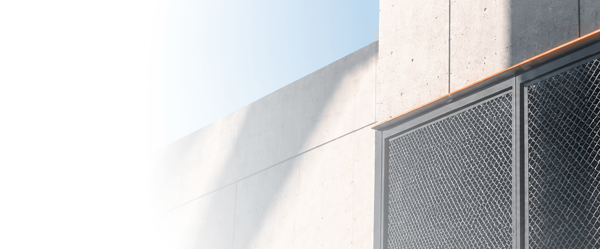 Sunlit concrete building facade with a copper trim line above a dark metal mesh panel